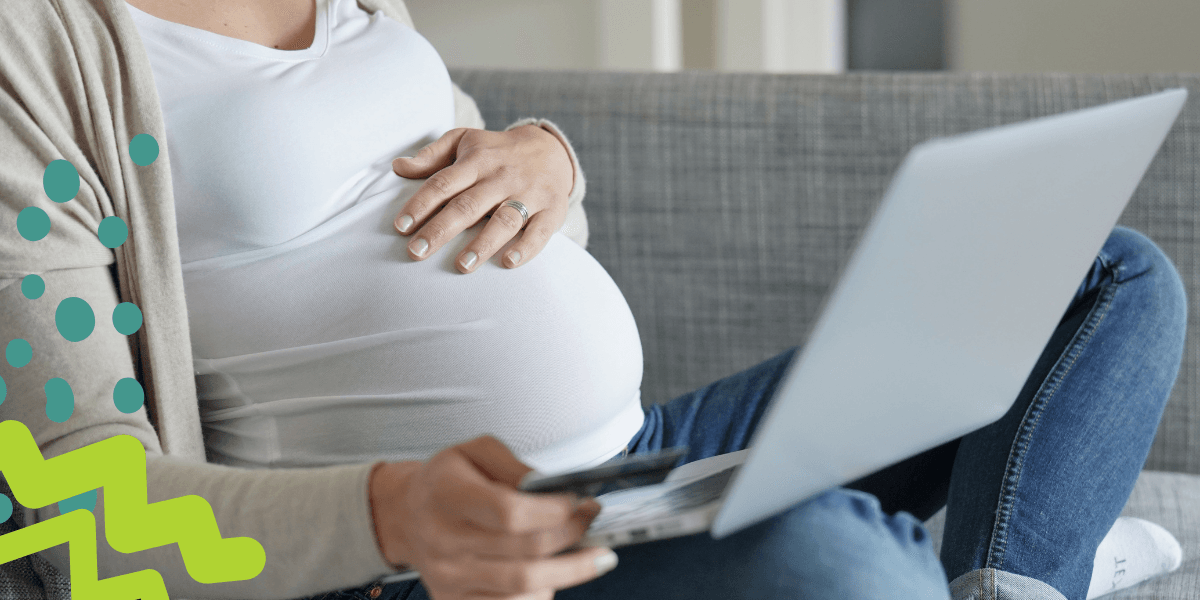 A pregnant woman sits cross-legged on her couch, hand on her belly, laptop in her lap.